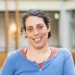 Jennifer Mankoff, a white, Jewish woman with an invisible disability. She is smiling broadly and standing casually in the Allen Center atrium