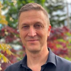 Mark Harniss a white man in his 50s with short brown hair and blue eyes wearing a dark polo shirt in front of fall-colored leaves.