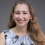 Kelly Mack, a young white woman with wavy brownish hair, smiles for the camera. She wears a blue floral blouse.